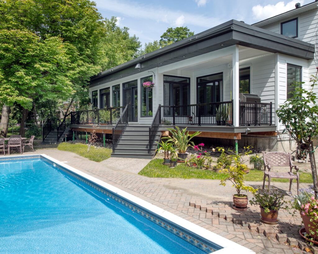 Modern white house with large windows and a covered porch overlooking a backyard patio and rectangular swimming pool, surrounded by trees and outdoor seating.