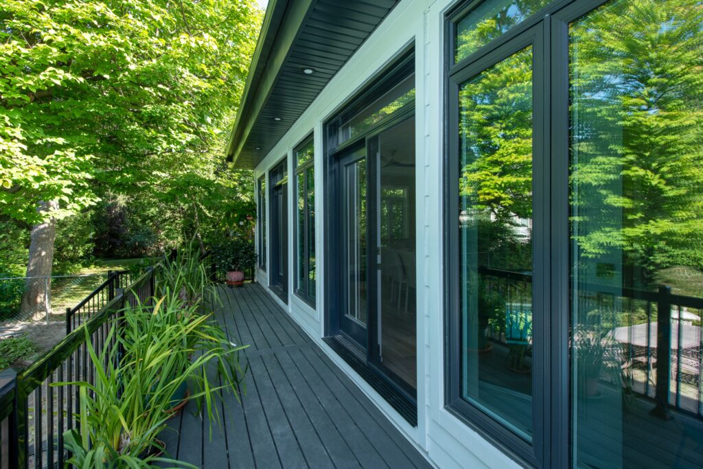 A modern house with large glass windows and a gray wooden deck, surrounded by lush green trees and potted plants, with black railings along the deck.