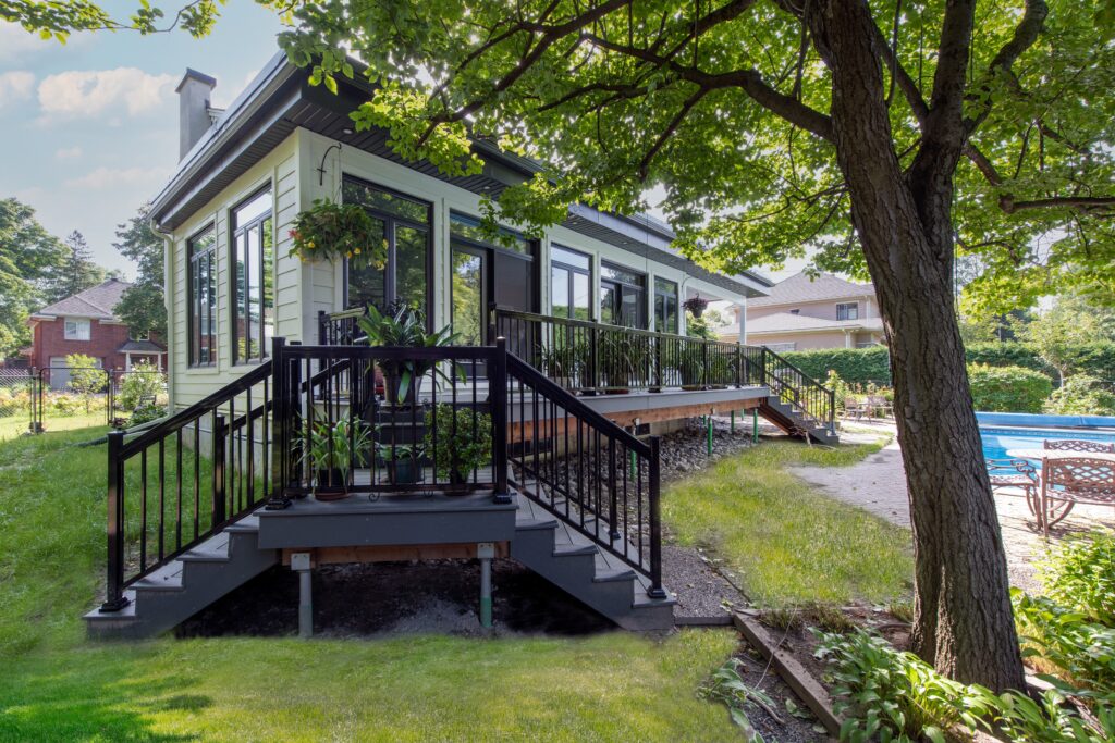 A modern house with large windows and a black-railed deck overlooks a grassy backyard with mature trees and an outdoor pool, under a bright, sunny sky.
