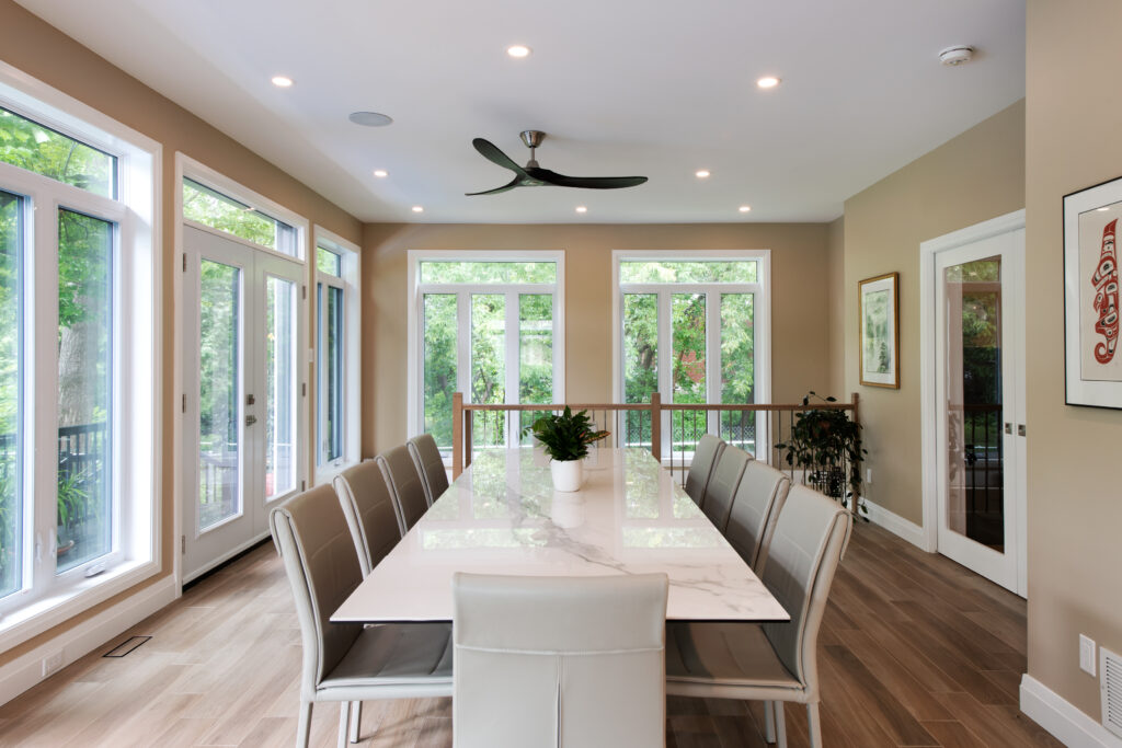 Modern dining room with a long marble table, ten beige chairs, large windows, glass doors, wood flooring, a ceiling fan, wall art, and a potted plant centerpiece. Lush greenery is visible outside.