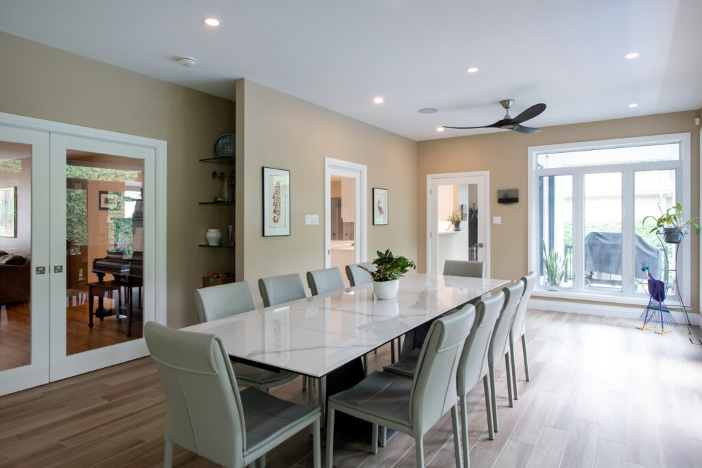 Modern dining room with a long white table, ten gray chairs, beige walls, large windows letting in natural light, plants, wall art, and a ceiling fan. Sliding doors lead to another room with a piano.