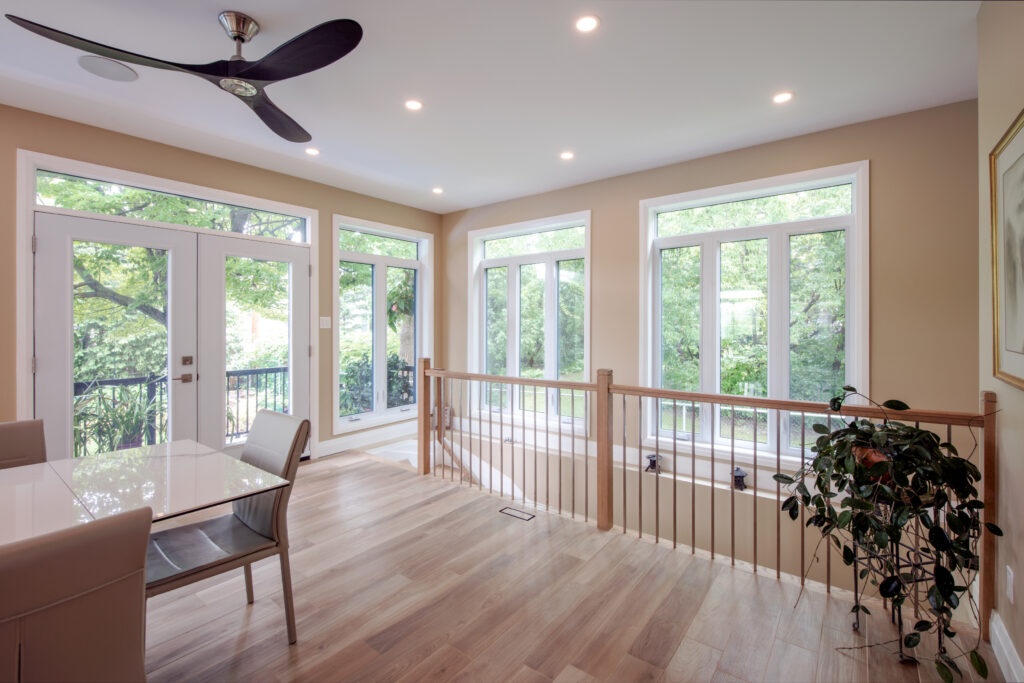 Bright, modern dining area with large windows, light wood floors, a glass table with white chairs, a ceiling fan, and a leafy plant near a wooden railing. Lush greenery is visible outside through the windows.