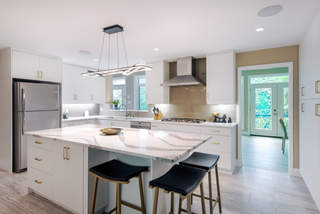 Modern kitchen with white cabinets, stainless steel appliances, marble island with black bar stools, geometric pendant light, and a view of greenery through glass doors in the background.
