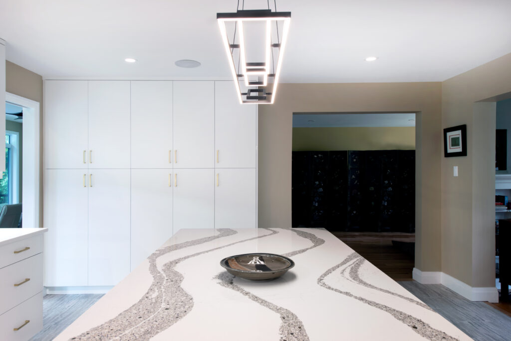 Modern kitchen with a large island featuring a white countertop with gray wave patterns, a decorative bowl in the center, white cabinets with gold handles, and a rectangular geometric pendant light above.