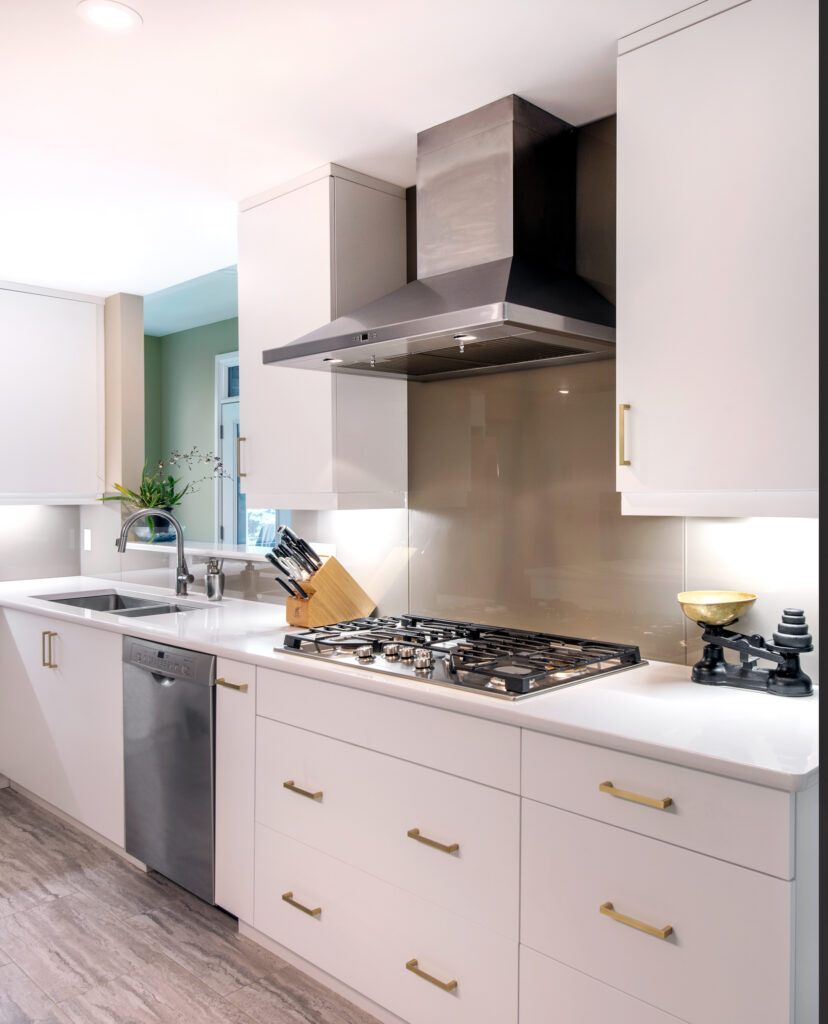 Modern kitchen with white cabinets, gold handles, a stainless steel gas stove and range hood, dishwasher, sink, knife block, and vintage kitchen scale on the countertop. Light wood flooring and under-cabinet lighting are visible.
