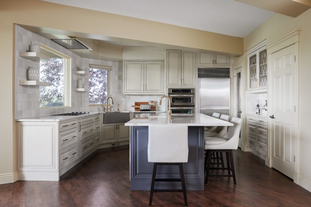 Modern kitchen with white cabinets, stainless steel appliances, a large island with a gray base, white countertop, and three bar stools. Hardwood floors and shelves with decor are also visible.