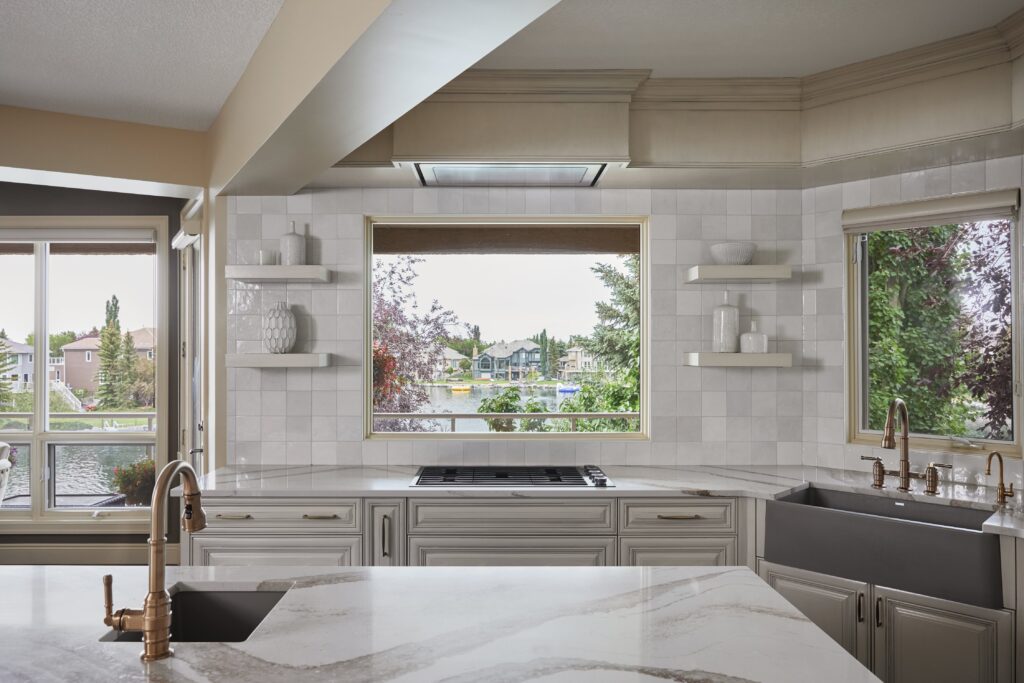Bright, modern kitchen with marble countertops, white cabinets, gold fixtures, and large windows overlooking a scenic suburban lake and trees. Open shelves and a farmhouse sink are visible under natural light.