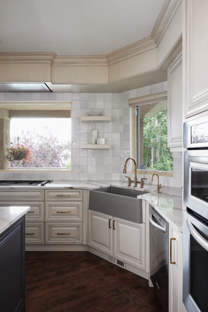 Modern kitchen with white cabinets, brass hardware, a large farmhouse sink, stainless steel double ovens, open shelves, and large windows showing greenery outside. Dark wood floors contrast with light countertops.