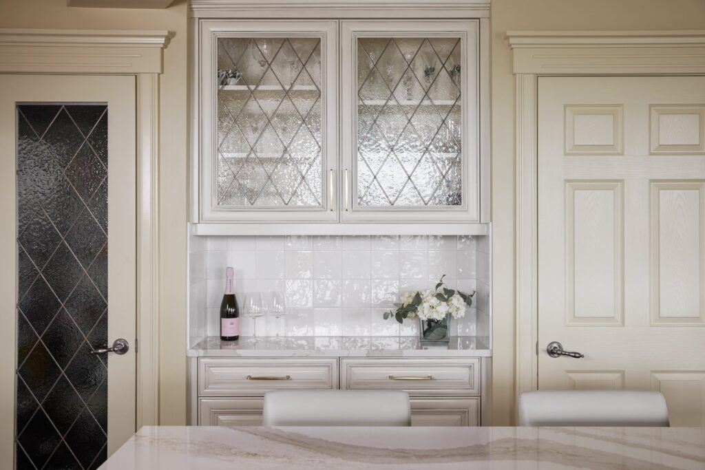 A bright kitchen features cream cabinets with glass doors, a marble countertop, a pink champagne bottle with glasses, and a vase of white flowers on the counter between two cream-colored doors.