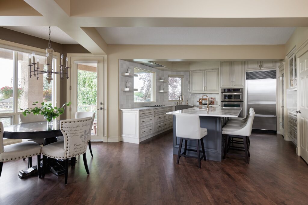 A modern kitchen with white cabinets, stainless steel appliances, a large island with bar stools, and a round dining table with upholstered chairs next to large windows.