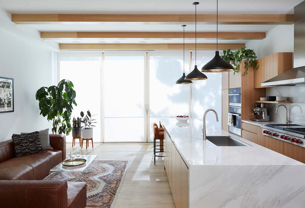 A modern kitchen with a marble island, hanging black pendant lights, built-in stainless steel appliances, a brown leather sofa, indoor plants, and large windows with sheer curtains letting in natural light.
