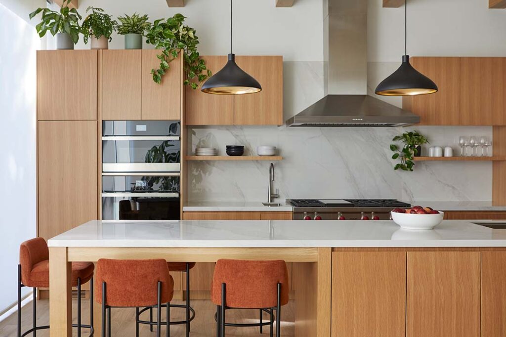 Modern kitchen with light wood cabinets, a white marble island, and orange upholstered bar stools. Black pendant lights hang over the island. Stainless steel appliances and green potted plants decorate the space.