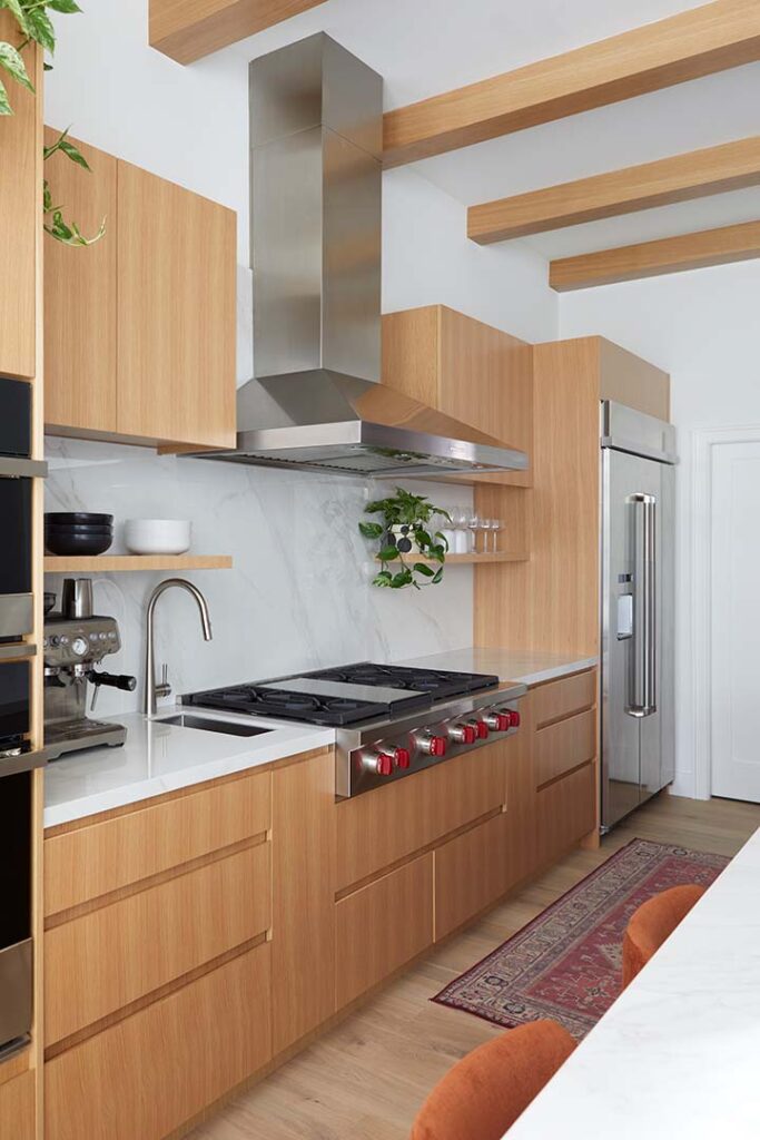 Modern kitchen with light wood cabinets, stainless steel appliances, a marble backsplash, and a red patterned rug. Open shelves hold dishes and a plant. Exposed wooden ceiling beams add warmth to the space.