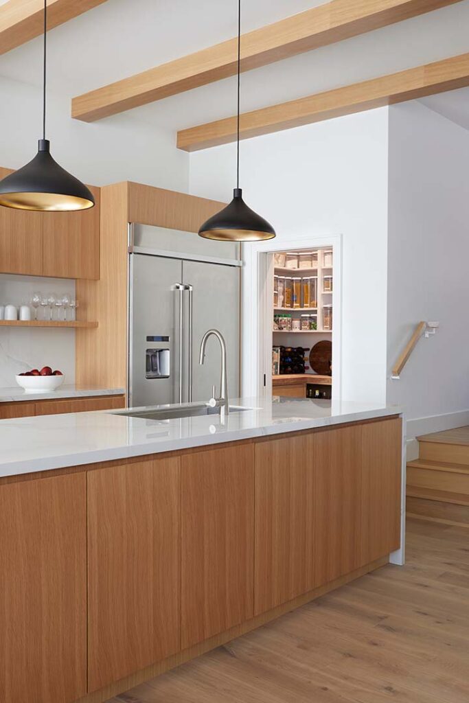 Modern kitchen with light wood cabinets, a white countertop, stainless steel refrigerator, pendant lights, open shelves with dishes, and a view into a pantry stocked with food items. Wooden ceiling beams are visible.