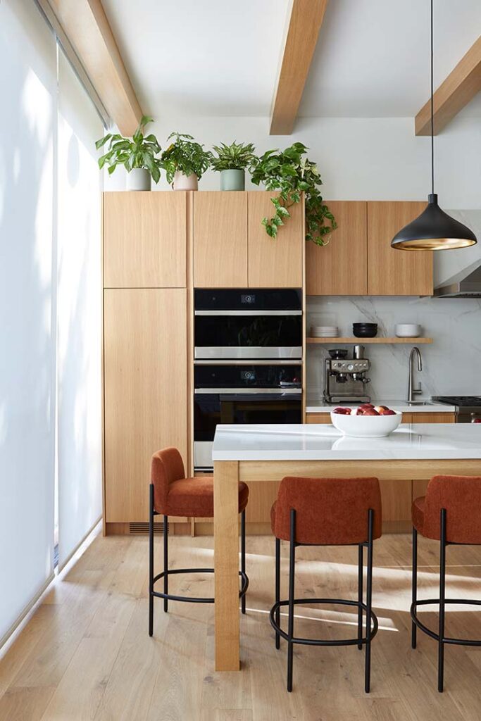 Modern kitchen with light wood cabinets, built-in appliances, a white countertop island with brown velvet barstools, potted plants on top of the cabinets, and a black pendant light hanging above.