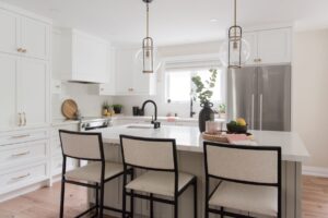 Modern kitchen with white cabinets, a large island with three cushioned stools, black fixtures, stainless steel fridge, pendant lights, and a vase with greenery on the island. Natural light enters through a window above the sink.