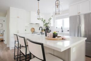 A modern white kitchen with a large island featuring three cushioned chairs, pendant lights, a black faucet, a vase with greenery, and a tray with fruit and cups; stainless steel appliances are visible.