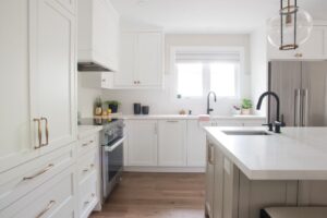Bright, modern kitchen with white cabinets, stainless steel appliances, a large island with a sink, light wood flooring, and minimalist decor, including potted plants and lemons on the countertop.