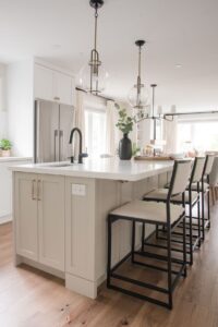 Modern kitchen with a large white island, beige cabinets, black faucet, and three high chairs. Pendant lights hang above the island, and a black vase with greenery sits on the countertop. Light wood floors and stainless steel appliances.