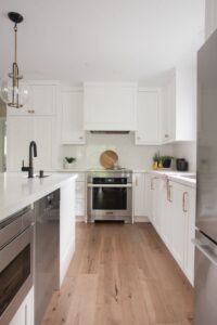 Bright, modern kitchen with white cabinets, light wood flooring, stainless steel appliances, a central island with a sink, and a stove with a cutting board and plant decor on the white backsplash.