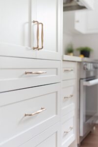 Close-up of white kitchen cabinets and drawers with brushed gold handles, next to a stainless steel stove and countertop with small green potted plants. The scene is bright and modern.