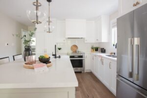 Modern kitchen with white cabinets, stainless steel appliances, a large island with a sink, pendant lights, and decorative items like a tray, plant, and bowl of fruit on the countertop. Light wood floor and bright natural lighting.