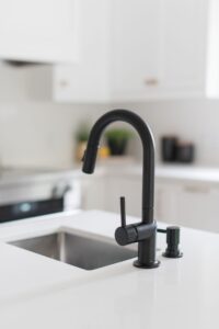 A matte black kitchen faucet with a curved spout is installed on a white countertop near a stainless steel sink in a modern, bright kitchen with white cabinets.