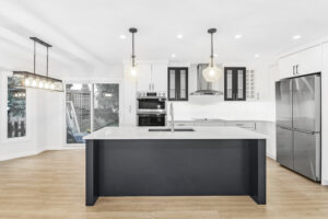Modern kitchen with a large island, pendant lights, stainless steel appliances, white cabinets, wood flooring, and a sliding glass door leading to an outdoor area.