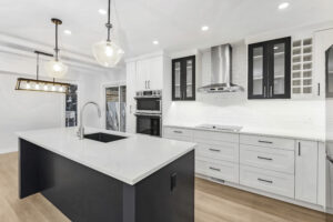Modern kitchen with white cabinets, a large island with built-in sink, stainless steel appliances, black accent features, pendant lights, light wood flooring, and a range hood over a white backsplash.
