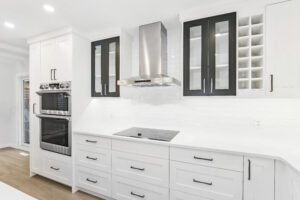 Modern kitchen with white cabinets, black-accented glass cupboard doors, stainless steel oven and hood, electric cooktop, wine rack, and white subway tile backsplash on a light wood floor.