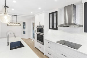 Modern white kitchen with stainless steel appliances, built-in oven, electric cooktop, and a large island with a sink. Bright lighting, wood flooring, and glass pendant lights enhance the clean, open space.