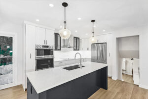 Modern kitchen with white cabinets, stainless steel appliances, a large island with a sink, pendant lights, and light wood floors. Glass doors open to an outdoor area, and stairs are visible beside the fridge.