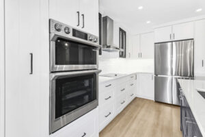 Modern kitchen with white cabinets, stainless steel double oven, refrigerator, and range hood, white countertops, and light wood flooring. The space is bright and clean with a minimalist design.