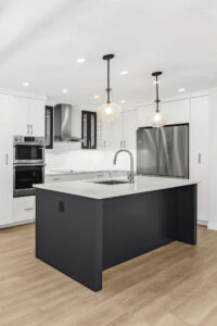 Modern kitchen with white cabinets, stainless steel appliances, a dark-colored island with a sink, pendant lights above, and light wood flooring. The space is bright and minimalistic.