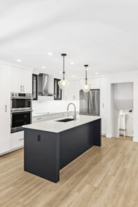 Modern kitchen with white cabinets, stainless steel appliances, a black island with a sink, pendant lights above, and light wood flooring. The space is bright and open with recessed ceiling lights.