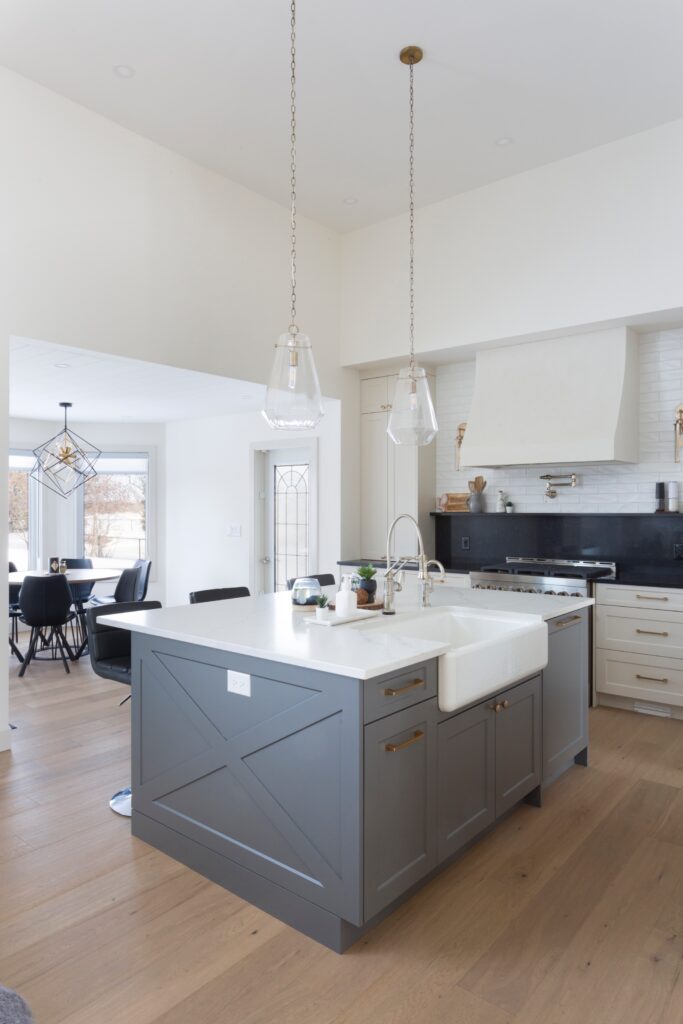 A modern kitchen with a gray island, white farmhouse sink, pendant lights, wood flooring, white cabinets, and stainless steel appliances. A dining area with a window is visible in the background.