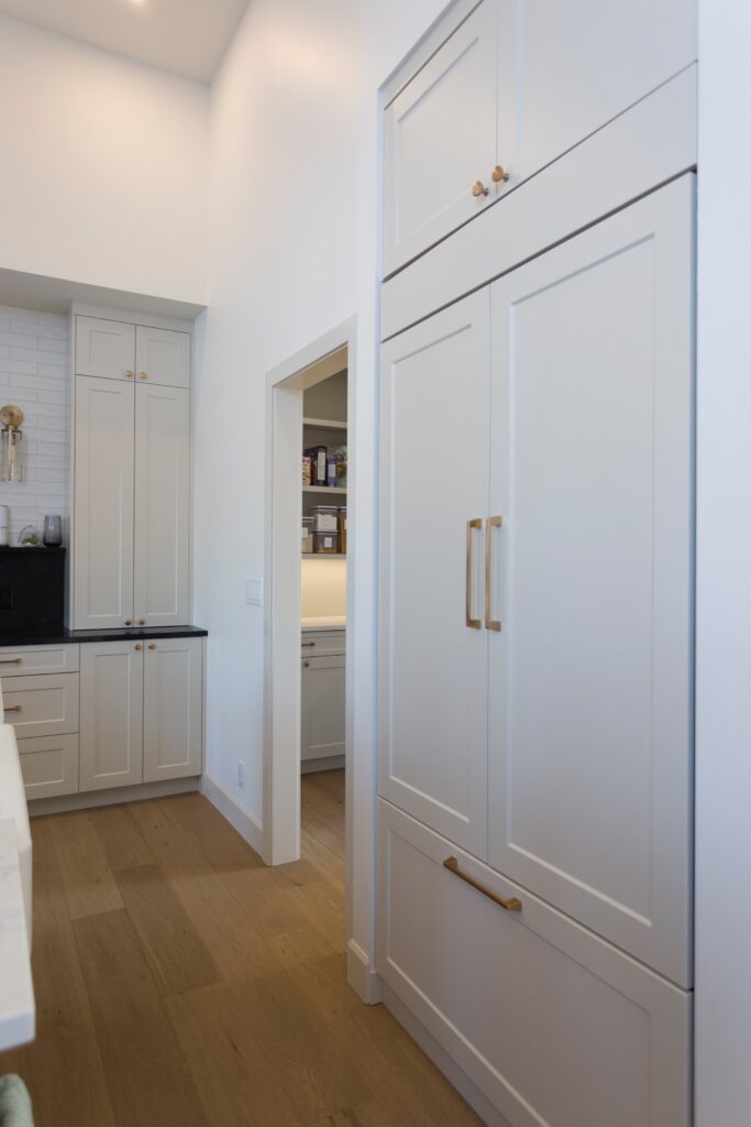 A modern kitchen with white cabinets, gold handles, light wood flooring, and a black countertop. There is a doorway leading to an adjacent room with built-in shelving.