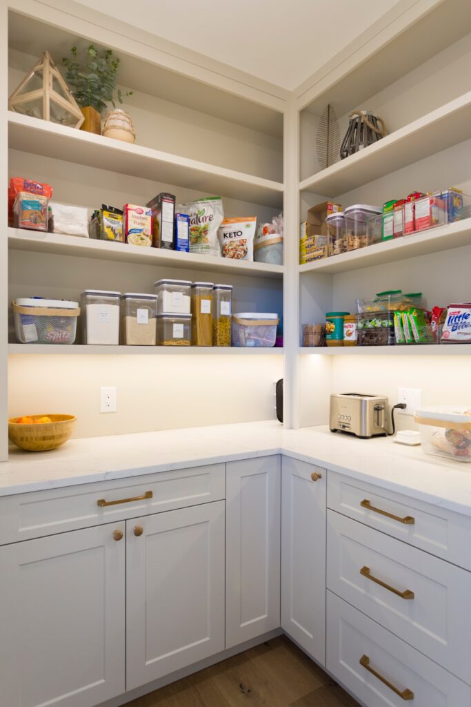 A neatly organized pantry with white shelves holding various food items, clear storage containers, cereal boxes, snacks, and kitchen appliances on a white counter with cabinets and drawers below.