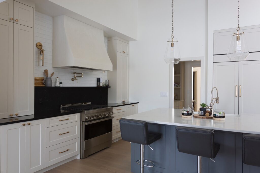 Modern kitchen with cream cabinets, a black countertop, stainless steel stove, and a range hood. An island with a white counter and two dark stools sits in front. Pendant lights hang above the island.