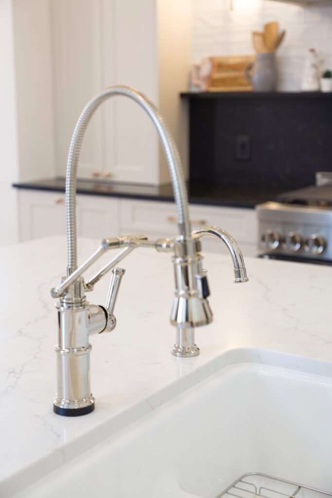 A close-up of a modern, shiny metal kitchen faucet with a flexible neck and lever handle, installed on a white marble countertop in a bright, contemporary kitchen.