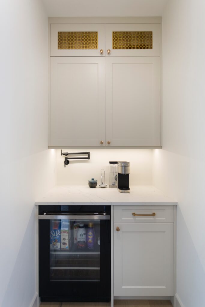 A small, modern kitchen nook with white cabinets, a built-in oven, a drawer, and a countertop featuring a coffee maker and a cup. Shelves behind the oven display drink cartons and bottles. Simple, clean design.