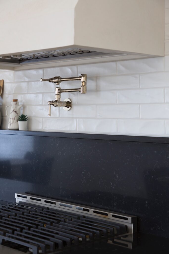 Modern kitchen with a white tile backsplash, a stainless steel pot filler faucet above a black stovetop, and a white range hood above. A small potted plant and decorative bottle sit on the counter.