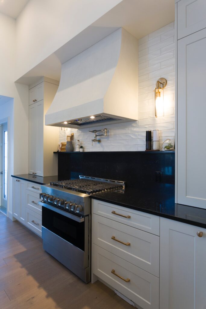 Modern kitchen with white cabinets, black countertops, stainless steel gas stove, large white range hood, white subway tile backsplash, brass handles, and a wall-mounted faucet above the stove. Warm lighting and wooden floor.