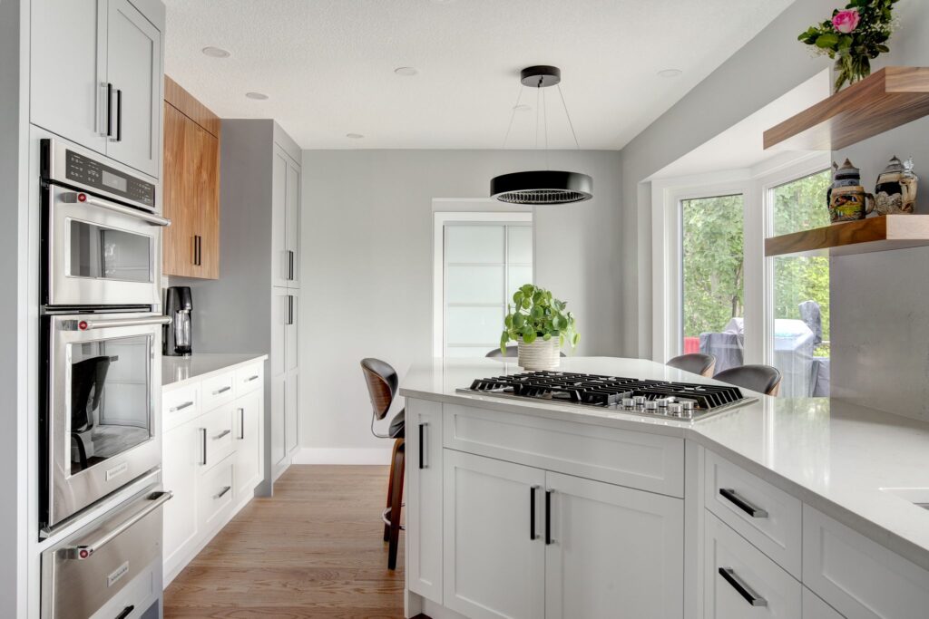 Modern kitchen with white cabinets, built-in ovens, a gas cooktop on a white island, wooden accents, floating shelves with decor, and large windows letting in natural light. A potted plant sits on the island.