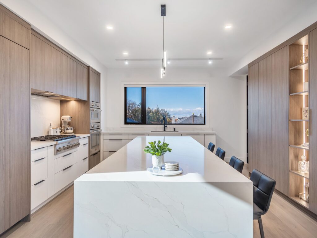 Modern kitchen with light wood cabinets, a large white marble island, black bar stools, built-in appliances, and a wide window with a view of trees and blue sky. A small plant sits on the island countertop.