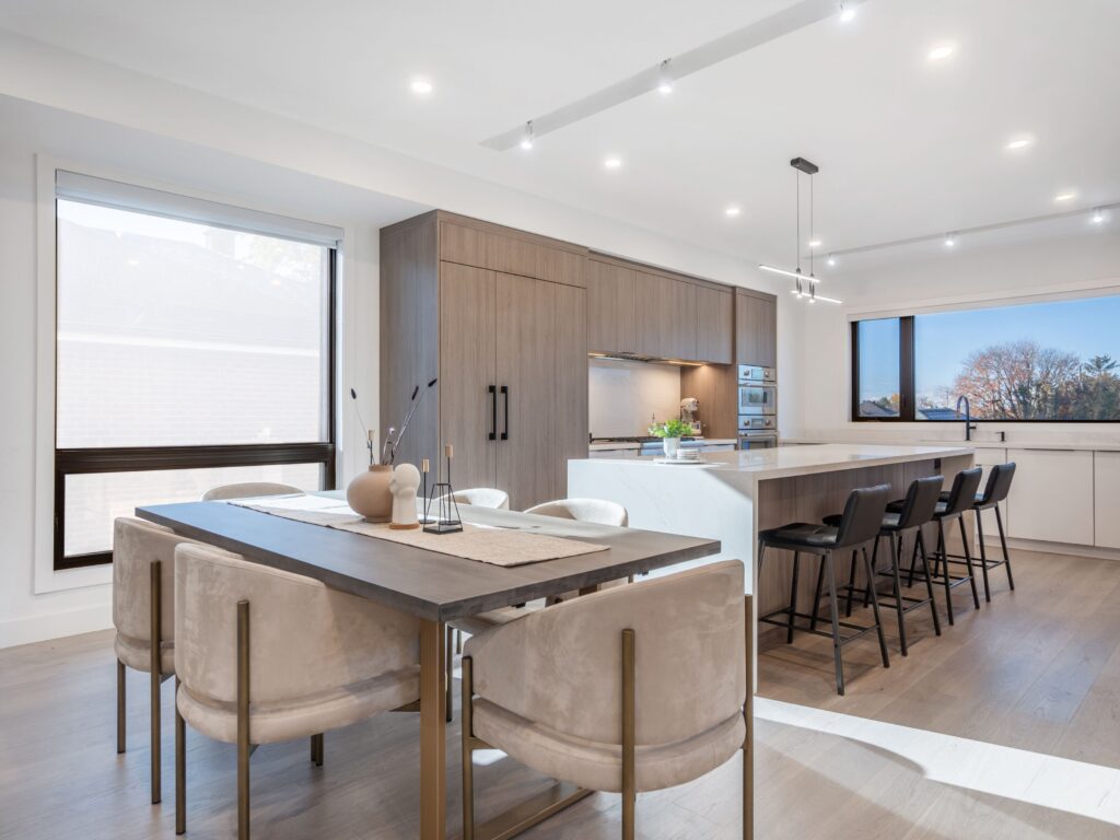 Modern kitchen and dining area with light wood floors, a large wooden dining table, beige upholstered chairs, and a sleek kitchen island with black barstools. Large windows let in natural light.
