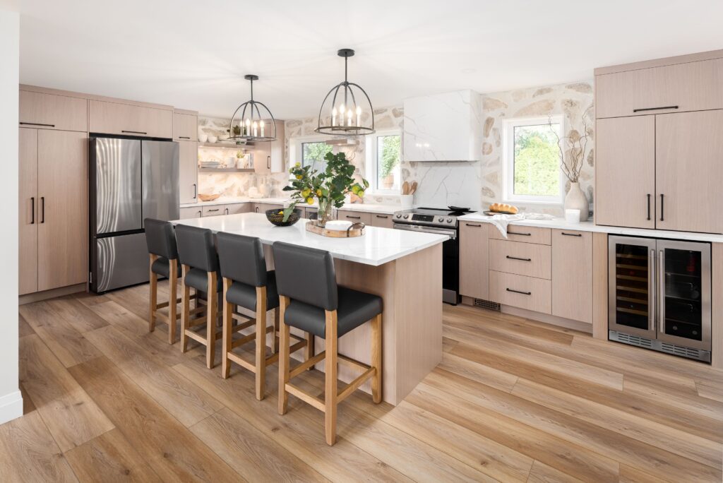 Modern kitchen with light wood cabinets, stainless steel appliances, a large island with four black barstools, pendant lights, wood flooring, and decorative plants and shelves. Natural light streams in through two windows.