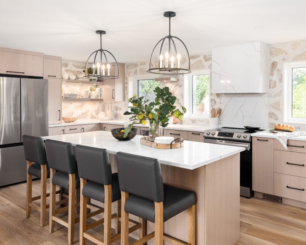 Modern kitchen with light wood cabinets, white marble countertops, stainless steel appliances, black pendant lights, and an island with four black barstools. A vase with greenery and lemons decorates the island.