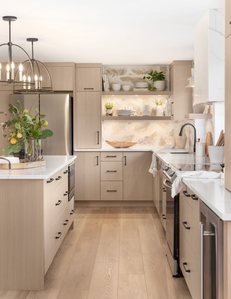 A modern kitchen with light wood cabinets, white countertops, stainless steel appliances, open shelves with dishes and plants, a marble backsplash, and a large island decorated with a vase of lemons.