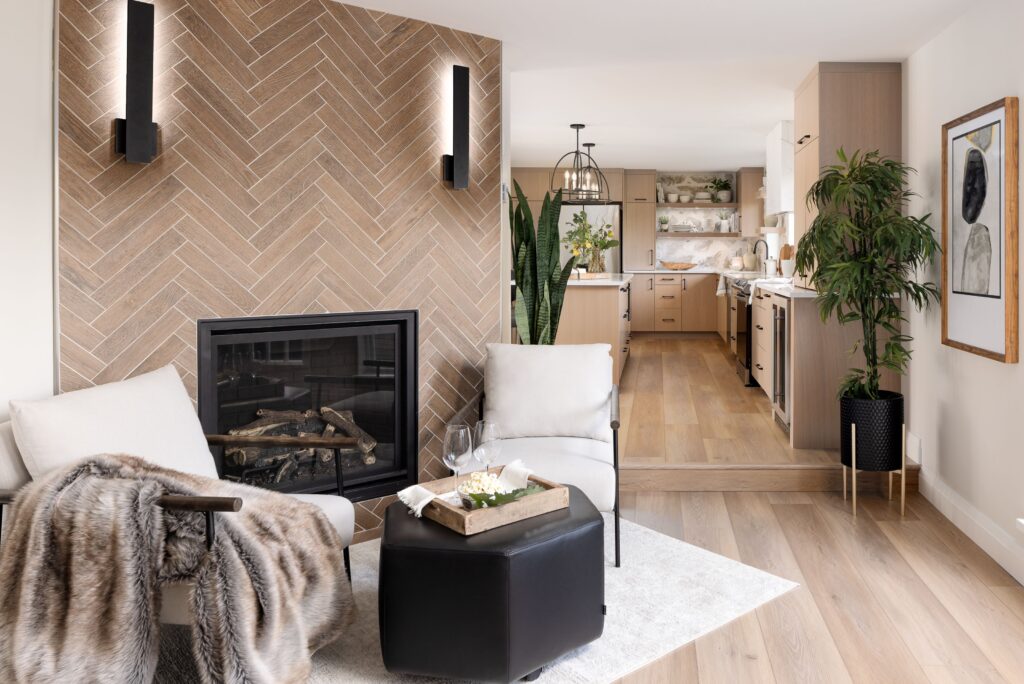 A modern living room with a chevron-patterned fireplace, two white chairs, a faux fur throw, a black ottoman with a tray, and a view into a light wood kitchen with green plants and framed art on the wall.
